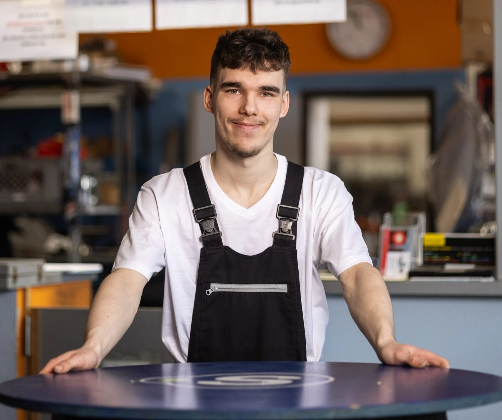 Ein Mann in Arbeitskleidung steht hinter einem runden Tisch, der auf einem Reifen montiert ist. Im Hintergrund sind Regale, Hinweisschilder und eine orangefarbene Wand zu sehen.