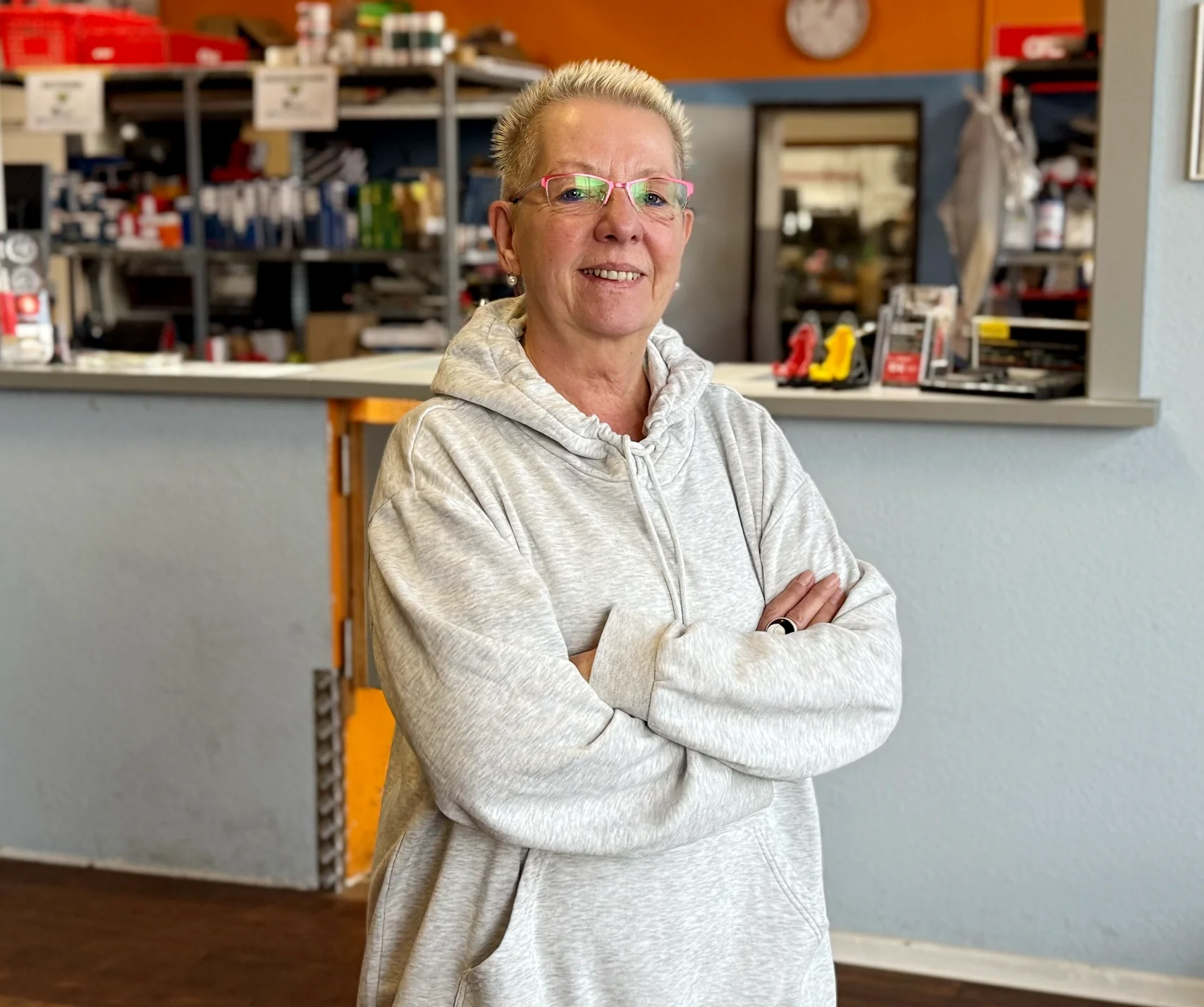 Eine Frau in Arbeitskleidung steht hin einer Werkstatt. Im Hintergrund sind Regale, Hinweisschilder und eine orangefarbene Wand zu sehen.